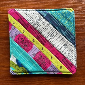 Puede incluir: Un posavasos de tela colorido con un diseño de patchwork que presenta rayas de tela azul, verde, amarilla, rosa y morada. La tela tiene un patrón impreso de notas musicales y rosas.