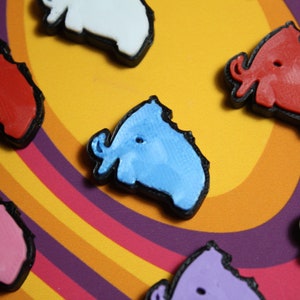 Peut inclure: Cinq figurines d'animaux colorées imprimées en 3D. Les figurines sont en forme d'éléphants et sont de différentes couleurs, notamment rouge, blanc, bleu et violet. Les figurines sont sur un fond multicolore.