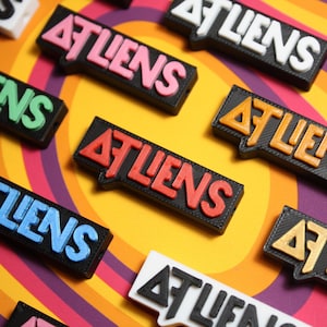 Könnte beinhalten: Eine Sammlung von bunten, rechteckigen, plastischen Namensschildern mit dem Wort "ATLiens" in verschiedenen Schriftarten und Farben darauf gedruckt. Die Schilder sind auf einem gelben und violetten gestreiften Hintergrund angeordnet.
