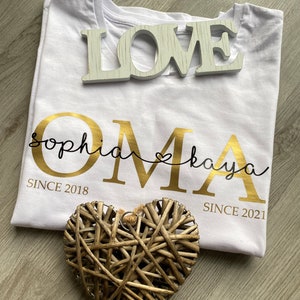 May include: A white t-shirt with gold lettering that reads "OMA" with the names "Sophia" and "Kaya" above and below the word "OMA". The shirt also has the dates "SINCE 2018" and "SINCE 2021". A wooden "LOVE" sign and a woven heart are also in the image.