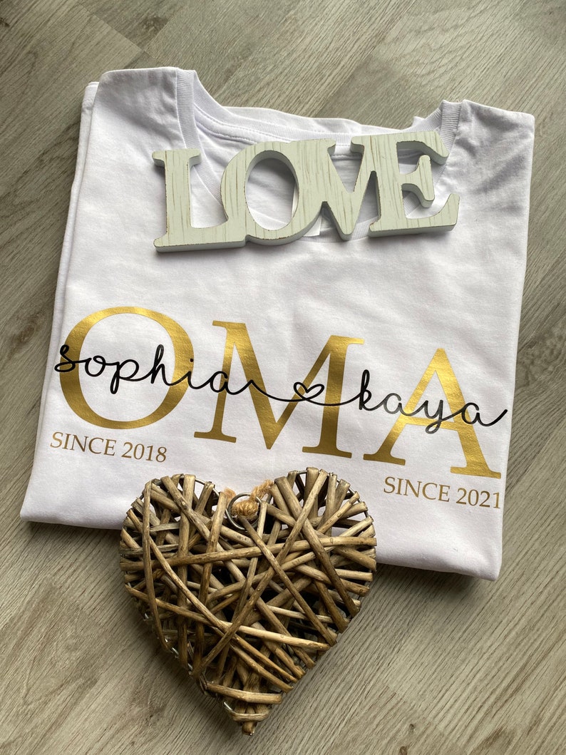 May include: A white t-shirt with gold lettering that reads "OMA" with the names "Sophia" and "Kaya" above and below the word "OMA". The shirt also has the dates "SINCE 2018" and "SINCE 2021". A wooden "LOVE" sign and a woven heart are also in the image.