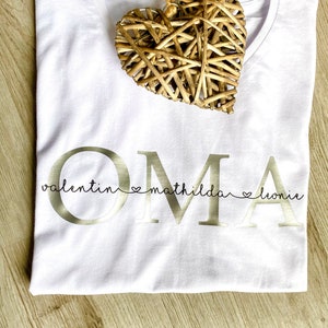 May include: White V-neck t-shirt with the word "OMA" in gold lettering, with the names "valentin mathilda leonie" below. A woven heart decoration rests on the shirt.