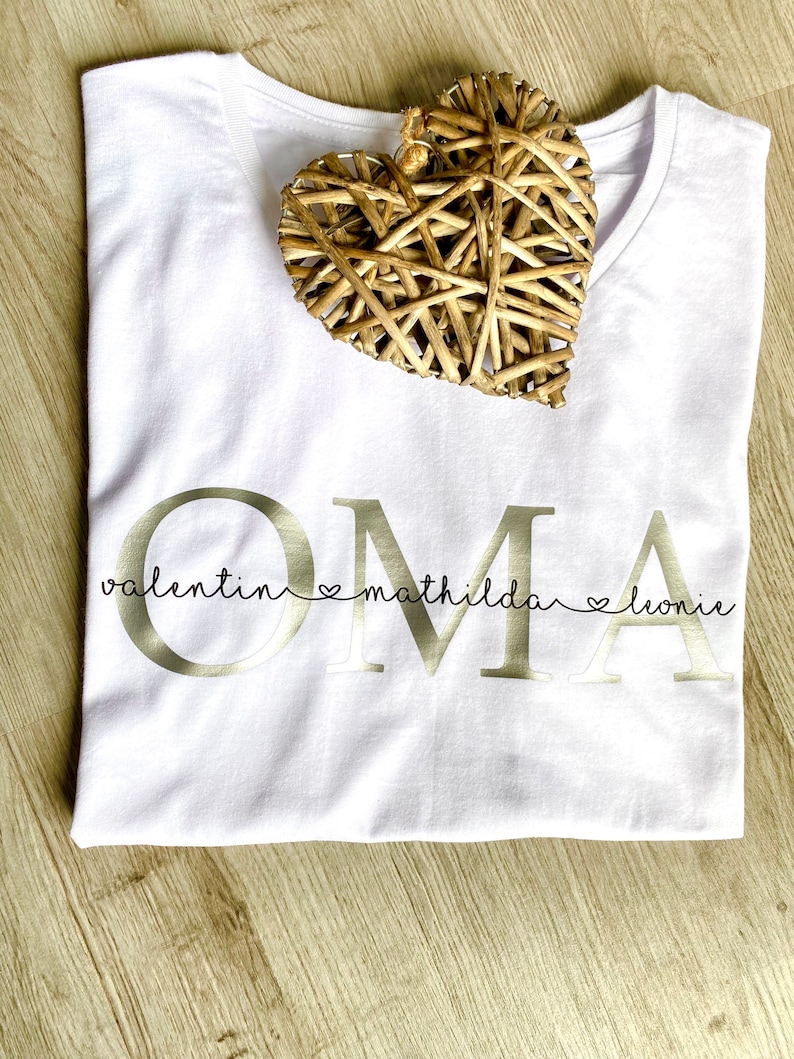 May include: White V-neck t-shirt with the word "OMA" in gold lettering, with the names "valentin mathilda leonie" below. A woven heart decoration rests on the shirt.