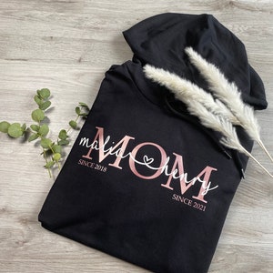 Könnte beinhalten: Schwarzer Kapuzenpullover mit dem Wort "MOM" in roségoldenen Buchstaben, mit den Namen "malia" und "henry" in weißer Schrift. Die Daten "SINCE 2018" und "SINCE 2021" sind ebenfalls abgebildet.