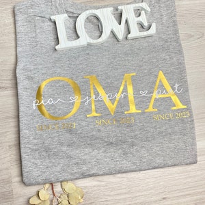 May include: A gray t-shirt with gold lettering that says "OMA" and the names "pia", "jasper", and "pit" underneath. The shirt also has the words "SINCE 2021" and "SINCE 2023". A white wooden "LOVE" sign rests on top of the shirt.