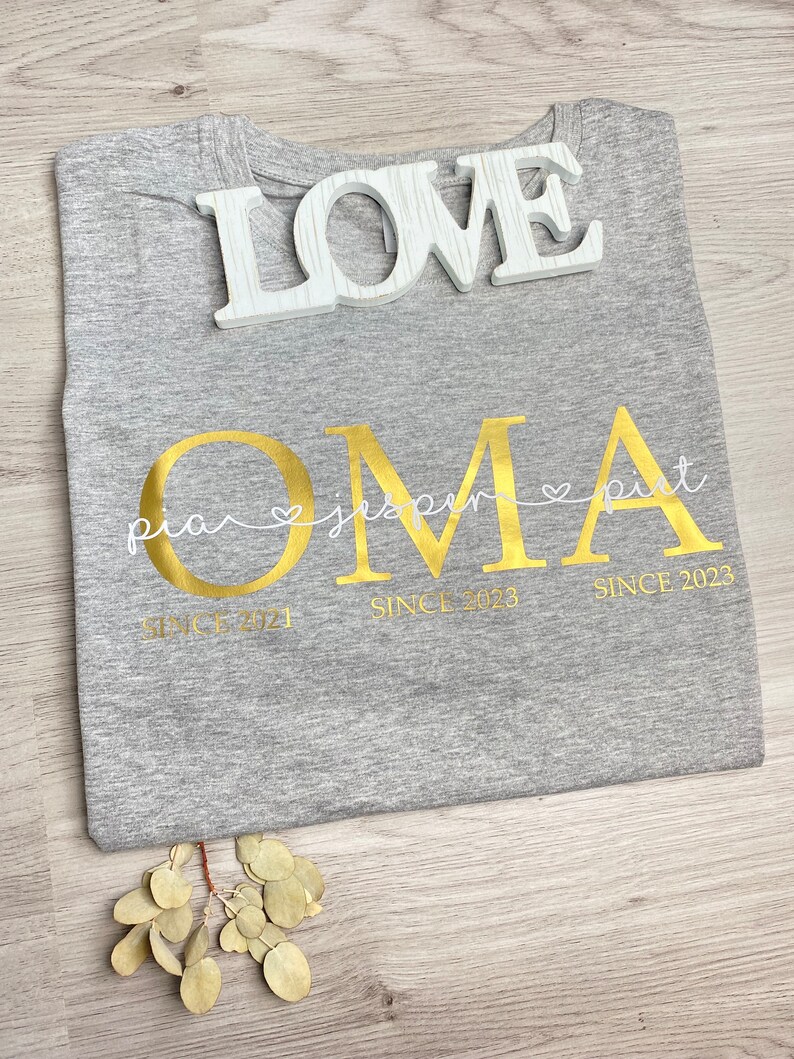 May include: A gray t-shirt with gold lettering that says "OMA" and the names "pia", "jasper", and "pit" underneath. The shirt also has the words "SINCE 2021" and "SINCE 2023". A white wooden "LOVE" sign rests on top of the shirt.
