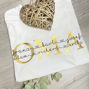 May include: A white t-shirt with the gold word "OMA" and names below it. The names are connected to the letters with hearts. A woven heart and eucalyptus sprig are on the shirt.
