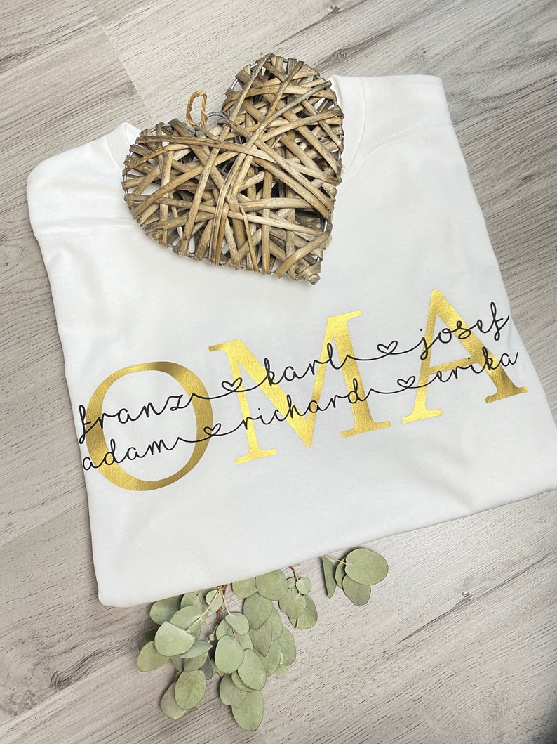 May include: A white t-shirt with the gold word "OMA" and names below it. The names are connected to the letters with hearts. A woven heart and eucalyptus sprig are on the shirt.