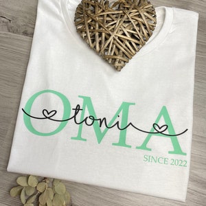 May include: White t-shirt with the word "OMA" in mint green, the name "toni" in a script font, and the text "SINCE 2022". A woven heart decoration rests on the shirt. The shirt is on a wooden surface.
