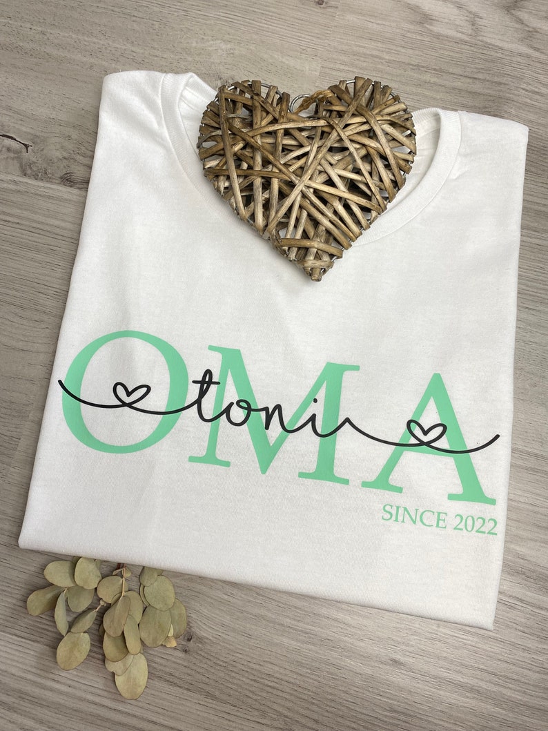 May include: White t-shirt with the word "OMA" in mint green, the name "toni" in a script font, and the text "SINCE 2022". A woven heart decoration rests on the shirt. The shirt is on a wooden surface.