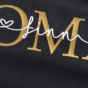 May include: Black fabric with the word "OMA" in gold, with the name "Finn" in a white script font. A white heart design is above the "O" and the "A". The fabric is slightly wrinkled.