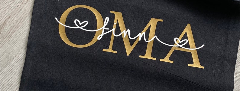May include: Black fabric with the word "OMA" in gold, with the name "Finn" in a white script font. A white heart design is above the "O" and the "A". The fabric is slightly wrinkled.