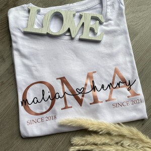 May include: White t-shirt with rose gold text that reads "OMA" with the names "malia" and "henry" above it, and the dates "SINCE 2018" and "SINCE 2021" below. A wooden "LOVE" sign sits on top of the shirt.