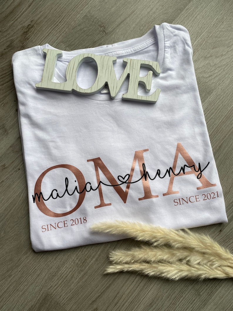 May include: White t-shirt with rose gold text that reads "OMA" with the names "malia" and "henry" above it, and the dates "SINCE 2018" and "SINCE 2021" below. A wooden "LOVE" sign sits on top of the shirt.