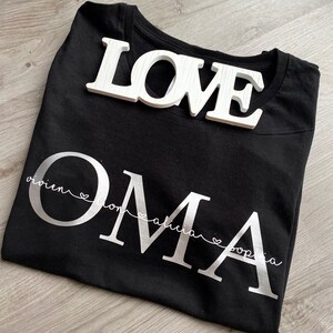 May include: Black t-shirt with the word "OMA" in large silver letters, with the names Vivien, Lona, Alicia, and Sophia underneath. A white wooden "LOVE" sign rests on top of the shirt.