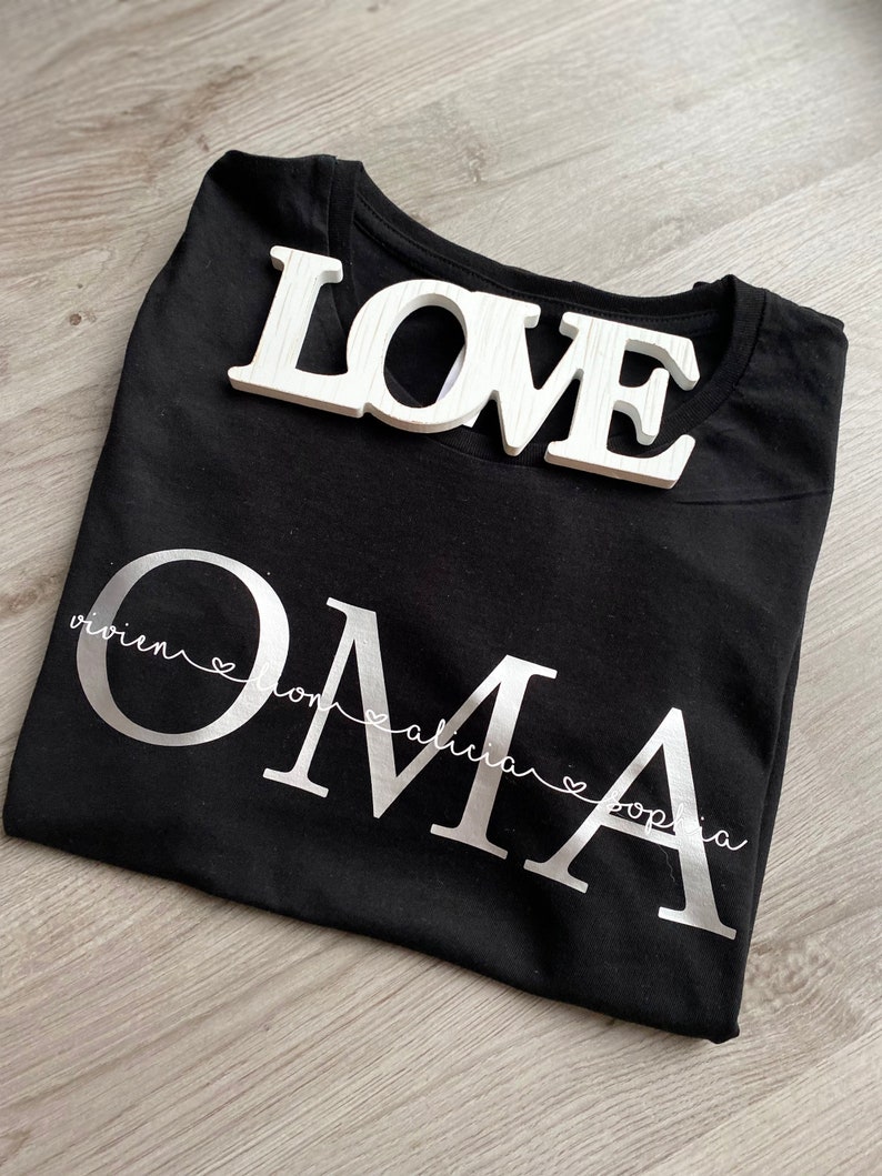 May include: Black t-shirt with the word "OMA" in large silver letters, with the names Vivien, Lona, Alicia, and Sophia underneath. A white wooden "LOVE" sign rests on top of the shirt.
