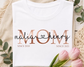May include: A white t-shirt featuring the word MOM in large rose gold letters. The names malia and henry are written in black cursive script across the center, connected by a small heart. The text SINCE 2018 and SINCE 2021 appears below.