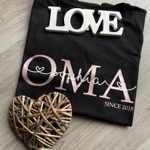May include: Black t-shirt with the word "Oma" in rose gold script, the name "Sophia" in white script, and "SINCE 2018". A white wooden "LOVE" sign and a woven heart are also present. The shirt is folded.