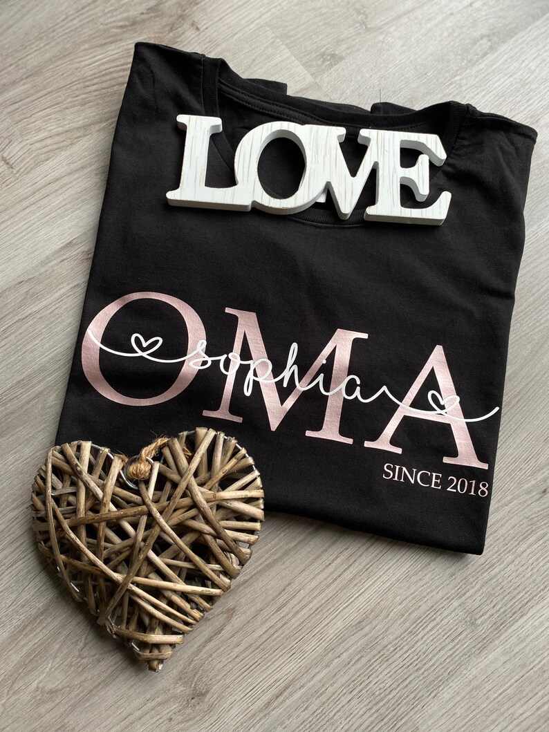 May include: Black t-shirt with the word "Oma" in rose gold script, the name "Sophia" in white script, and "SINCE 2018". A white wooden "LOVE" sign and a woven heart are also present. The shirt is folded.