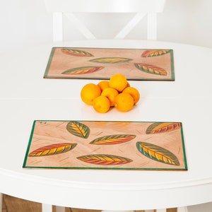 Puede incluir: Una mesa de comedor redonda blanca con dos manteles individuales rectangulares con diseños de hojas en tonos naranja, amarillo y verde. Un grupo de naranjas está en el centro. La mesa está rodeada de sillas blancas.