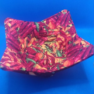 May include: A red and yellow fabric bowl with a chili pepper pattern. The bowl is folded and sitting on a blue surface.