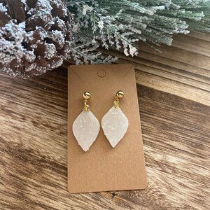 Könnte beinhalten: Ein Paar weiße glitzernde Ohrringe mit goldenen Akzenten. Die Ohrringe sind tropfenförmig und haben oben eine kleine goldene Kugel.