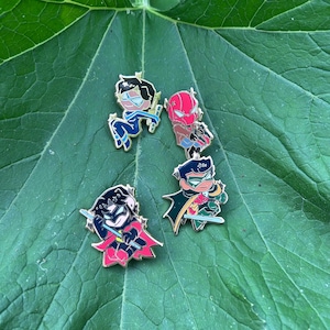 May include: Four enamel pins featuring stylised cartoon characters. One character wears a blue suit with a mask, one wears a red helmet and brown jacket, one wears a black and red suit with a cape, and one wears a green and red suit with a cape.