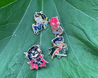 May include: Four enamel pins featuring stylized cartoon characters. One character wears a blue suit with a mask, one wears a red helmet and brown jacket, one wears a black and red suit with a cape, and one wears a green and red suit with a cape.
