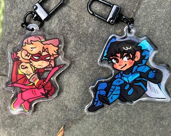 May include: Two acrylic keychains featuring cartoon characters. One keychain depicts a character in red and pink with a bow and arrow. The other keychain shows a character in blue and black. Both keychains have black metal clasps.
