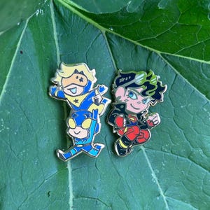 Puede incluir: Dos pines de esmalte con personajes de dibujos animados. Uno muestra a un personaje rubio con traje azul y amarillo sobre los hombros de una figura azul. El segundo pin representa a un personaje de pelo oscuro con chaqueta negra y pantalones rojos.