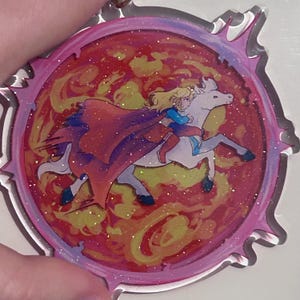 May include: A circular acrylic charm with a cartoon illustration of a person riding a white horse. The rider wears a blue top and a red cape. The background is a swirling mix of red, orange, and yellow, with glitter accents. The charm has a pink border.