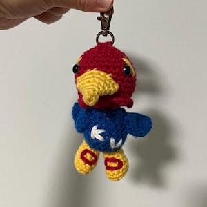 May include: A crocheted bird keychain with a red head, yellow beak, blue body, and white accents. The bird is attached to a metal ring.