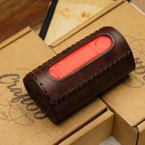 May include: A brown leather case with a red interior rests on two cardboard boxes. The case has stitched detailing and a rectangular opening. The boxes have the text "Craft66 Handmade Est. 2016" printed on them.
