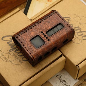 Puede incluir: Una funda de cuero marrón con un intrincado diseño floral, con dos ventanas de visualización rectangulares y varios agujeros pequeños. La funda se coloca encima de dos cajas de cartón con el logotipo "Craft66 Handmade".