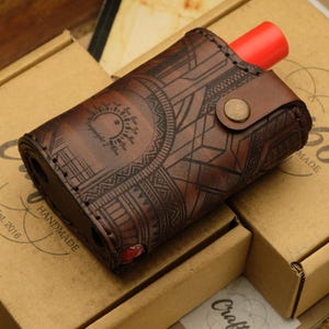 May include: A brown leather case with detailed geometric and sunburst patterns, holding a red cylindrical item. The case is fastened with a button. It sits on two cardboard boxes.