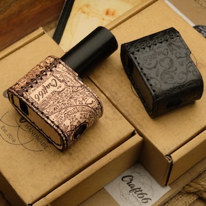 May include: Two leather cases, one tan with an intricate design and the text "Craft66", and one black with a floral pattern, sit on cardboard boxes. A black cylindrical object is next to the tan case. The boxes have the text "Est. 2016 HANDMADE".