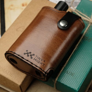 May include: A brown leather flask with a black strap and a silver button closure. The flask has the words "Tiny Might" and a small logo printed on it.