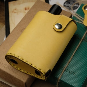 May include: A yellow leather flask with a metal closure and a strap. The flask is handmade and is sitting on a brown box with the word "HANDMADE" printed on it.
