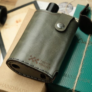 Peut inclure: Un étui en cuir vert avec un bord cousu brun et une fermeture à pression métallique. L'étui porte l'inscription "Tiny Might" et un logo imprimé. L'étui est posé sur une boîte brune avec le mot "Handmade" imprimé dessus.