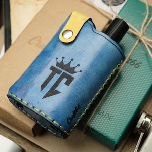Peut inclure: Un étui en cuir bleu avec une sangle jaune et un logo de couronne. L'étui est destiné à une petite bouteille de parfum ou de cologne. L'étui est fait à la main et porte le texte "Craft 66" sur le côté.