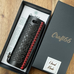 Puede incluir: Funda de teléfono de cuero negro con un diseño de nudo celta cosido en rojo. La funda está en una caja negra con el texto "Craft66" y "Hand Made" en ella.