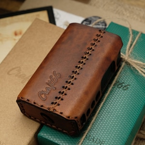 Puede incluir: Una funda de cuero marrón con la palabra "Craft66" grabada en la parte delantera. La funda tiene un diseño cosido y está sobre una caja verde con una caja marrón detrás.