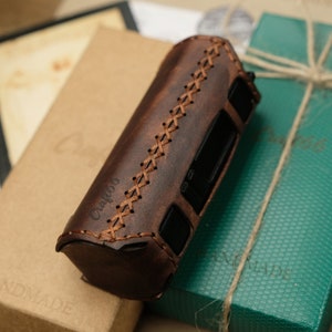 Puede incluir: Una funda de cuero marrón con costuras y la palabra "Craftbox" grabada en el lateral. La funda está sobre una caja verde con una cuerda marrón atada alrededor.