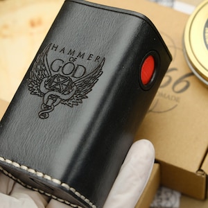 Könnte beinhalten: Schwarze Lederbox mit weißem Stepprand. Die Box hat einen roten Knopf an der Seite und ist mit den Worten "HAMMER OF GOD" und einem geflügelten Schlangenmotiv graviert.