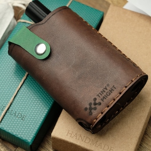 May include: A brown leather case with a green strap and a silver button. The case has the text "TINY MIGHT" and "HANDMADE" printed on it.