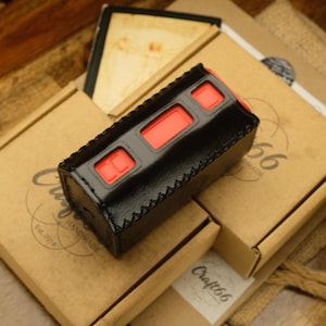 May include: A black leather case with red detailing, possibly for a vaping device, sits on a stack of cardboard boxes. The case features three rectangular openings and a stitched edge. The boxes display a circular logo and the text "Craft66 Handmade Est. 2016".