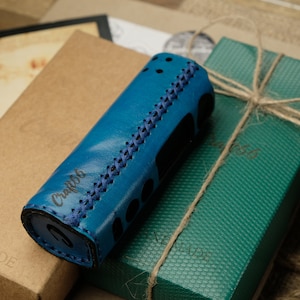 Puede incluir: Una funda de cuero color turquesa con costuras azules, posiblemente para un dispositivo de vapeo, descansa sobre una caja marrón y una caja de regalo verde atada con hilo. La funda tiene el texto "Craft56".