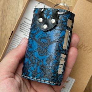 Puede incluir: Un llavero de cuero azul con un diseño de calavera y flores. El llavero tiene un lazo de metal para sujetar las llaves y un cierre de presión.