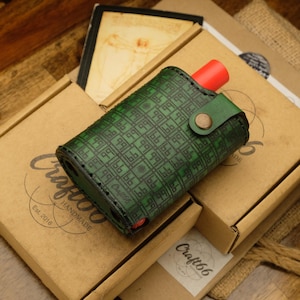 Peut inclure: Étui en cuir vert avec un motif géométrique, un composant cylindrique rouge et une sangle verte fixée par un bouton. L'étui repose sur une boîte en carton avec le logo "Craft66" et le texte "HANDMADE Est. 2016".