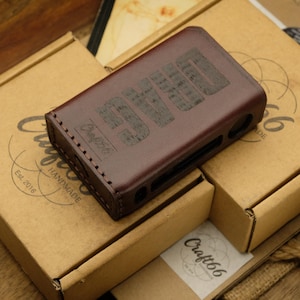 Puede incluir: Una funda de cuero marrón con la palabra "SMD" grabada descansa sobre dos cajas de cartón. Las cajas tienen el logotipo "Craft66 Handmade". La funda es rectangular con bordes redondeados y recortes.
